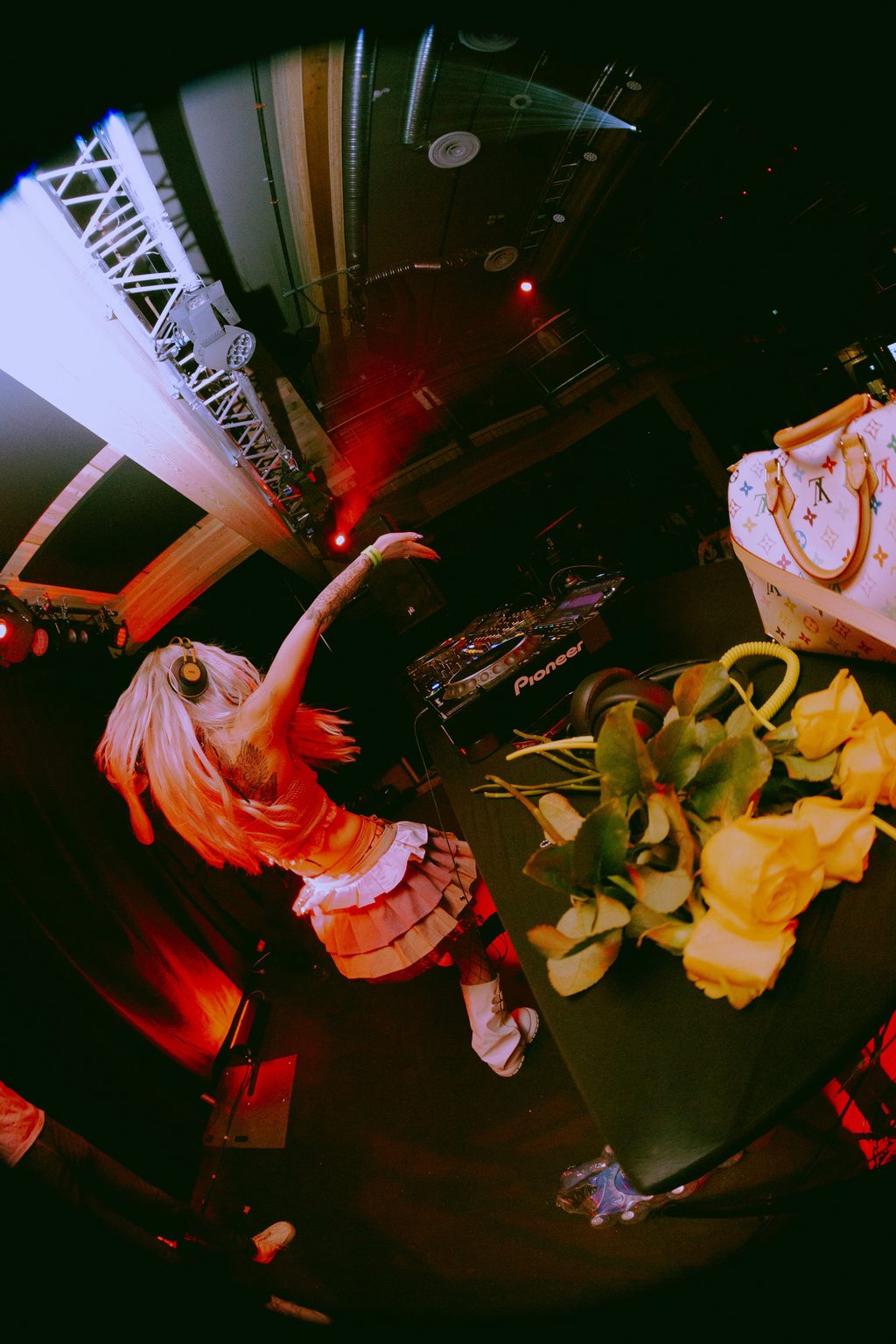 Fisheye shot of ROZENZA DJing with arm raised at the decks, yellow roses on the table under red club lighting