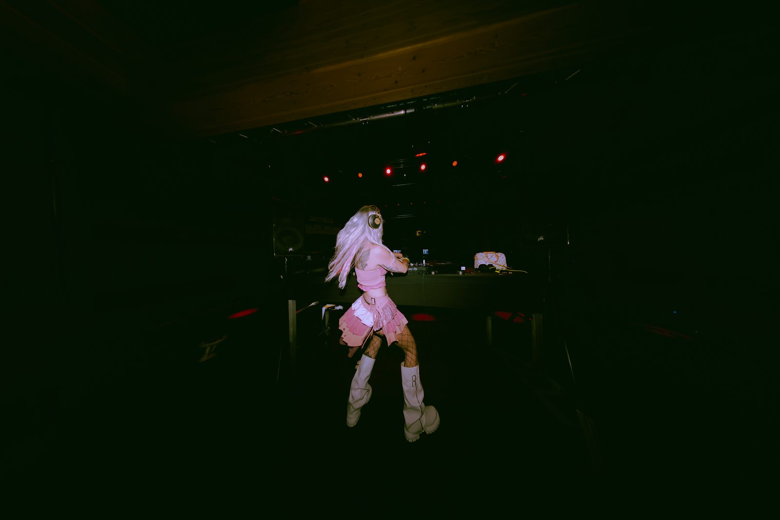 ROZENZA seen from behind at the DJ decks in a dark venue, wearing a pink outfit and white boots under red stage lights