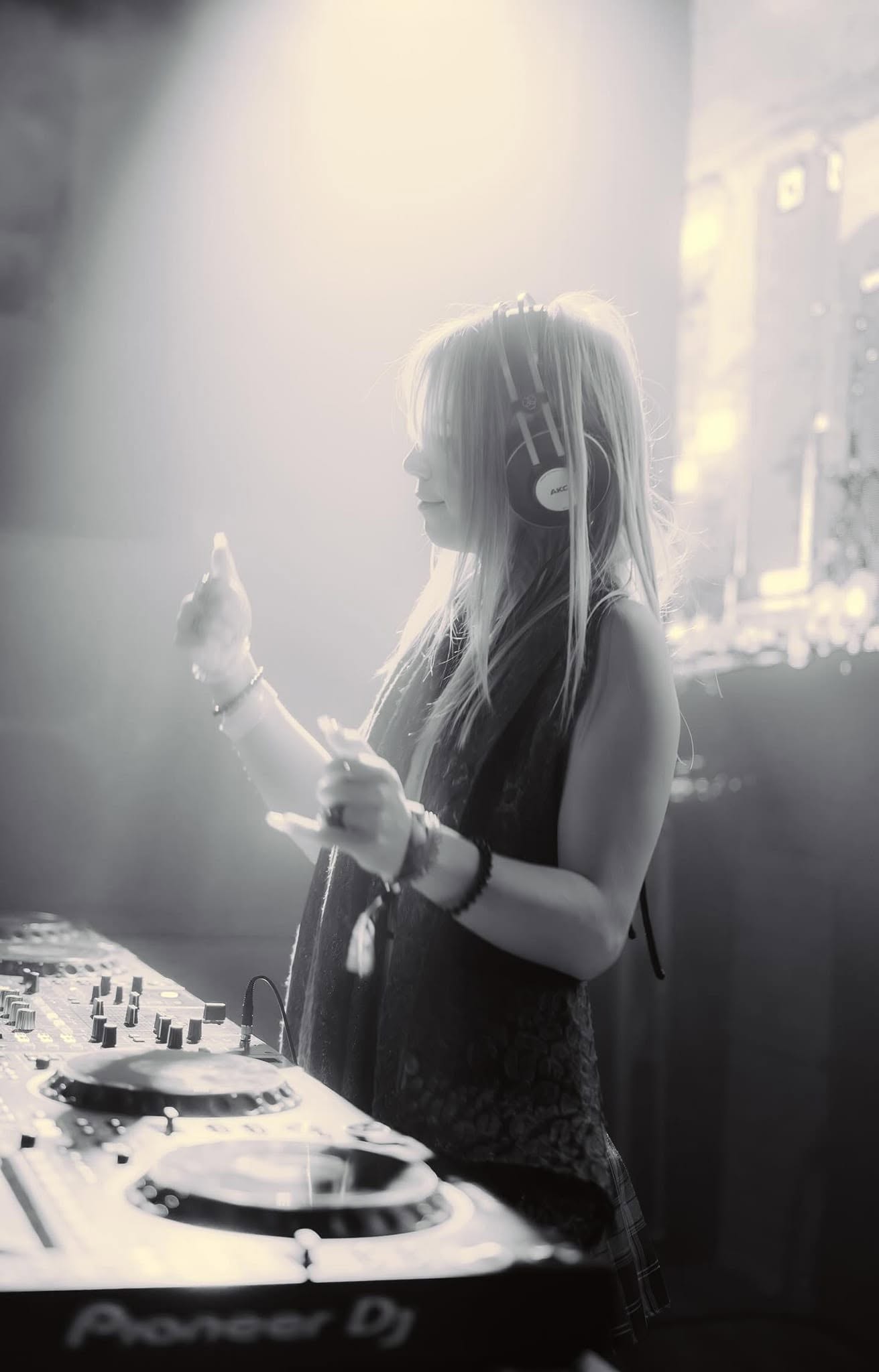 ROZENZA in profile at the decks wearing headphones, gesturing toward the crowd